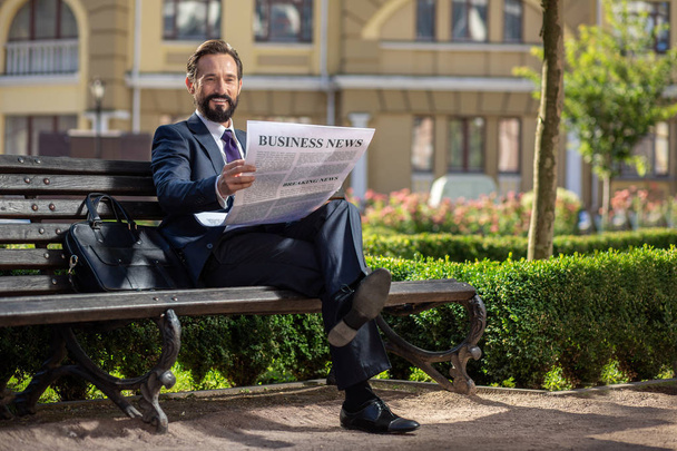 ビジネス動向。ベンチに座りながら新聞を読む笑顔の自信に満ちたビジネスマンの全長 - 写真・画像