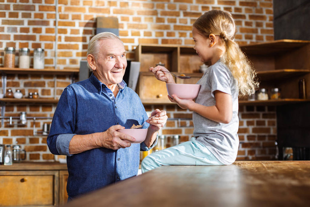 Healthy food. Positive elderly man eating cereal while enjoying time with his granddaughter - Foto, imagen