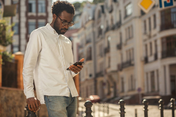Concentrated young African American man thoughtfully looking at the screen of his smartphone while standing in the street - Фото, изображение