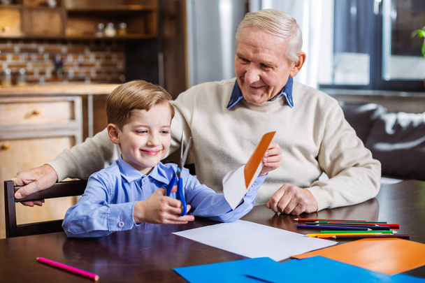 Creative work. Cheerful little boy making cutouts while making a festive card with his grandfather - Foto, immagini