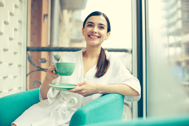 Happy woman in the armchair holding cup and smiling - 写真・画像