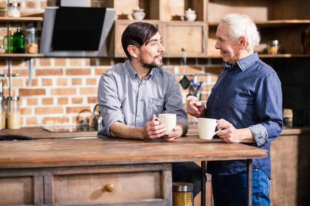 Tasty beverage. Cheerful handsome man drinking coffee with his father while standing in the kitchen - Фото, зображення