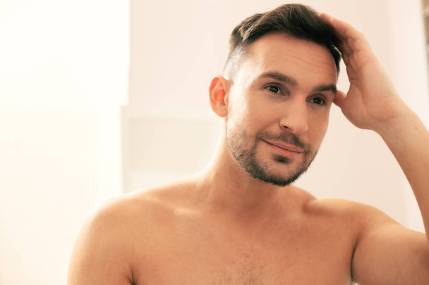 Attractive young man with bare shoulders looking confident and smiling while touching his hair - Foto, afbeelding