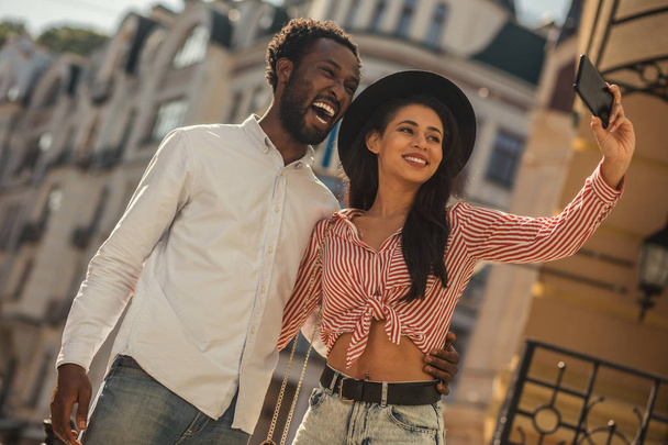 Excited young man laughing with his mouth open while young lady in a hat taking selfie with him - Foto, Imagen