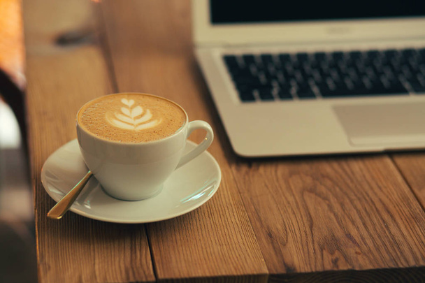 Selective focus of a white cup of coffee placed on a plate. Modern laptop on the background behind the cup - Foto, Bild