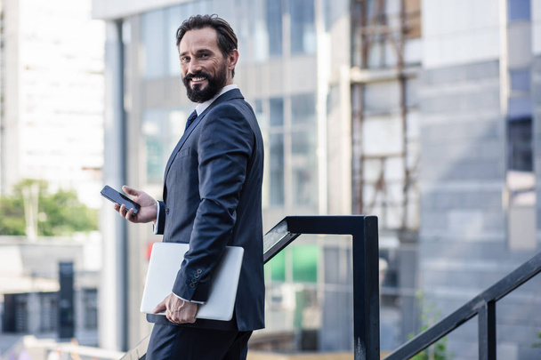 Well-dressed. Waist up of a positive bearded businessman holding his laptop while using smartphone - Foto, Bild