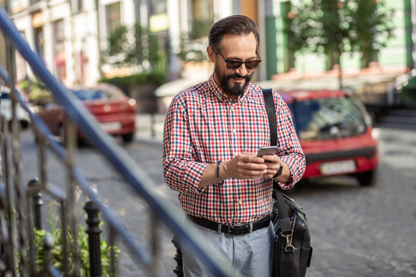 Internet user. Positive smiling man walking along the street while using his smartphone - Foto, immagini