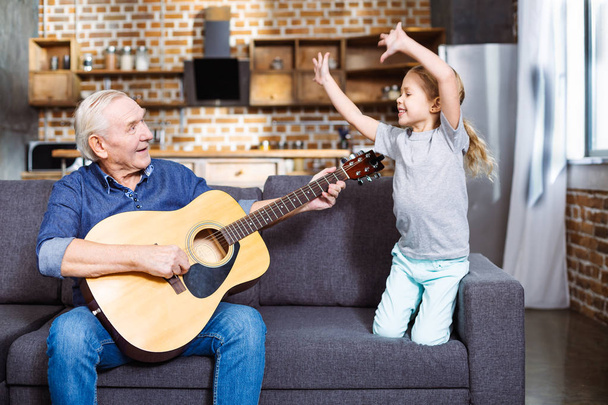 Active movements. Cheerful aged man playing the guitar while his granddaughter having fun on the couch - Photo, Image
