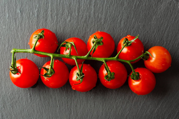Horizontal image of the fresh branch of ripe red cherry tomatoes lying on the black slate surface - Foto, afbeelding
