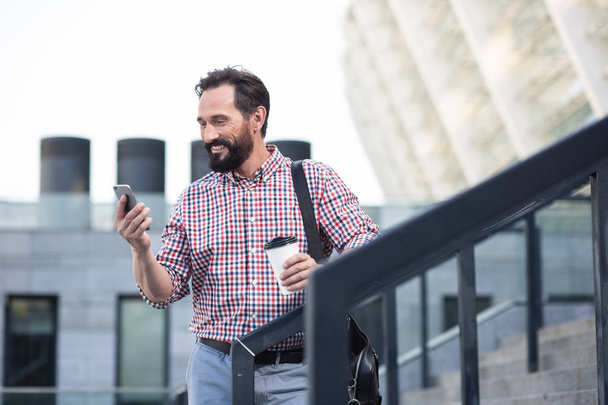 Street lifestyle. Cheerful handsome bearded man using his phone while standing outdoors - Foto, Imagem