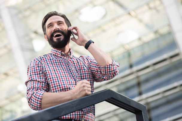 Glimlachend knappe man staande in stedelijke instellingen tijdens het praten op de telefoon - Foto, afbeelding