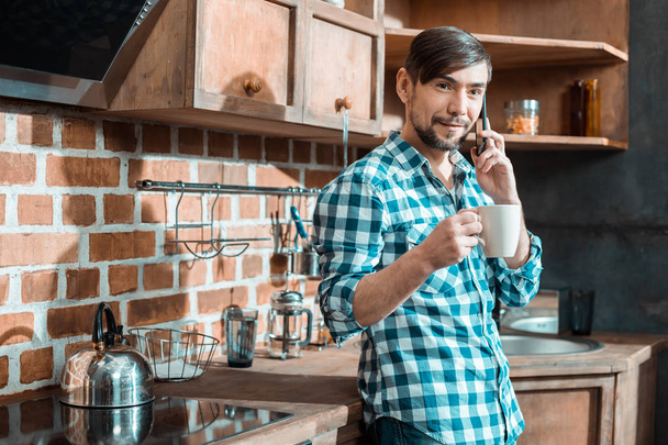 Mobile network. Smart nice bearded man standing in the kitchen and holding a cup of tea while making a call - Zdjęcie, obraz