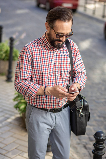 Digital lifestyle. Pleasant delighted man standing in the street while typing a message - Photo, Image