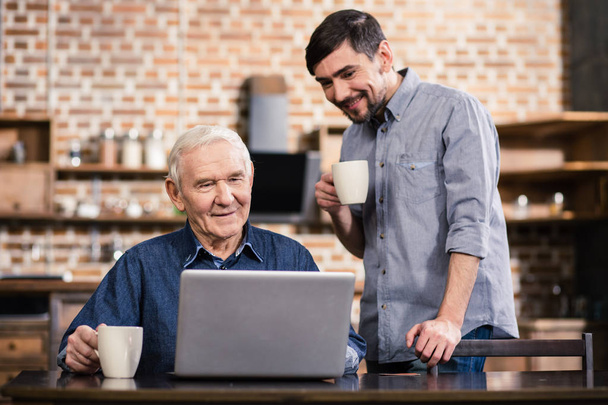 Modern user. Positive elderly man using his laptop while his son standing nearby - 写真・画像