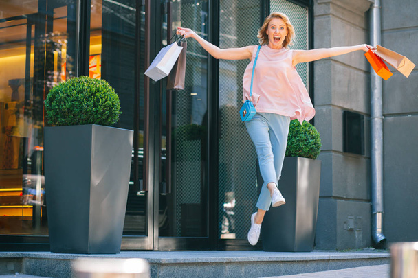 Захоплена жінка з паперовими сумками стрибає біля магазину і посміхається
 - Фото, зображення