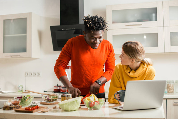 Young man looking attentively at the screen of modern laptop while cutting vegetables with his curious girlfriend standing near - Photo, image