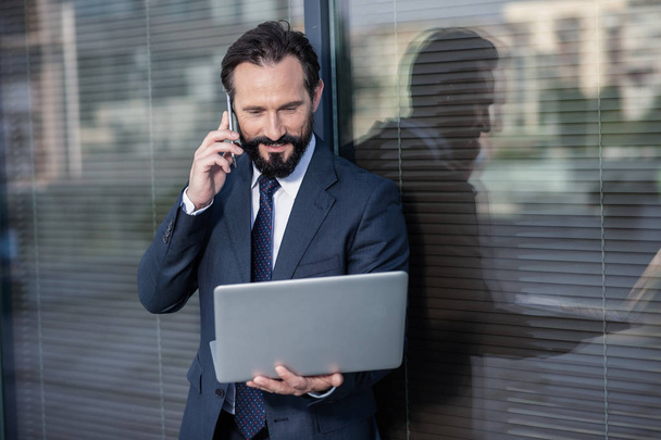 Digital world. Waist up of a pleasant professional businessman using laptop while talking on phone - Фото, изображение