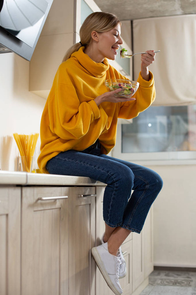 Cheerful relaxed woman on the kitchen countertop eating salad from the glass bowl and smiling - Фото, изображение
