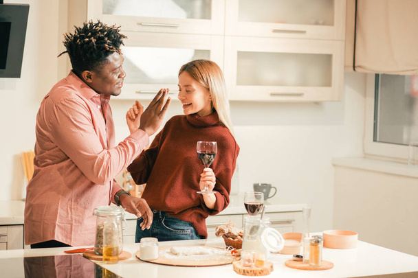 Waist up of positive young lady standing in front of her boyfriend in the kitchen and holding a glass of wine wheil having flour spot on the nose - Foto, Imagen
