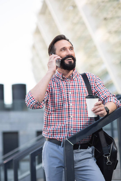 Full of joy. Cheerful pleasant man standing outdoors while having a conversation on phone - Foto, Imagen