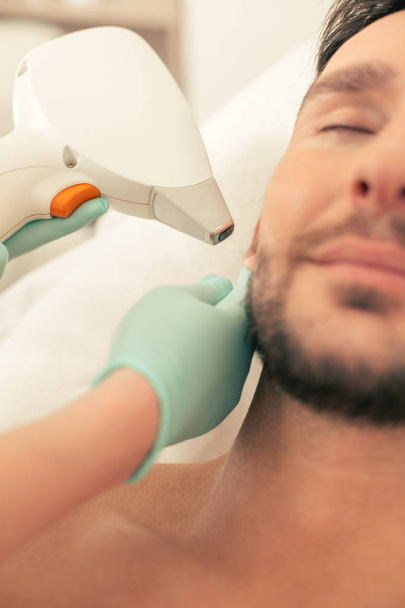 Close up photo of the bearded man with closed eyes and hands in rubber gloves conducting the laser hair removal and touching the ear of man - Foto, imagen