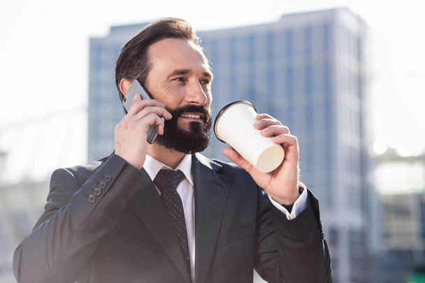 Call me back. Close up of a confident smiling businessman talking on phone while drinking morning coffee - Foto, Bild
