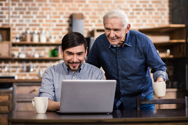 Just interested. Cheerful smiling man uisng laptop while his father feeling curious - Фото, изображение
