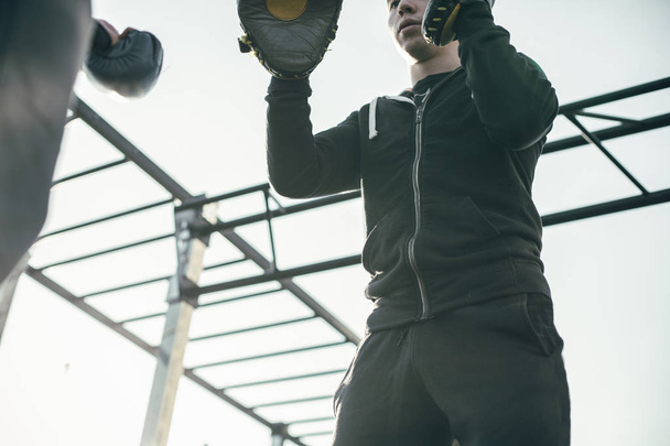 Confident young man in sports clothes wearing focus mitts during the boxing training - Φωτογραφία, εικόνα