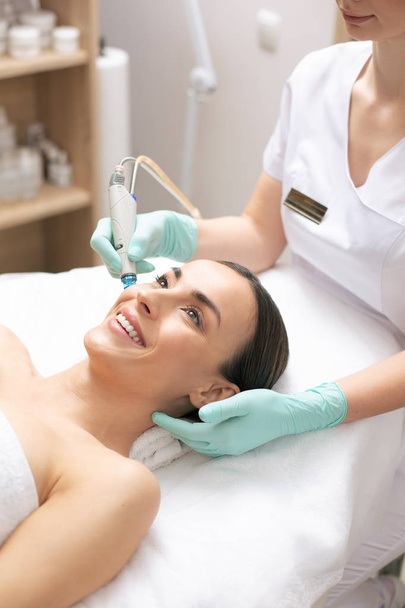 Smiling young woman enjoying the process of skin nourishment in modern clinic with a help of a special tool - Φωτογραφία, εικόνα