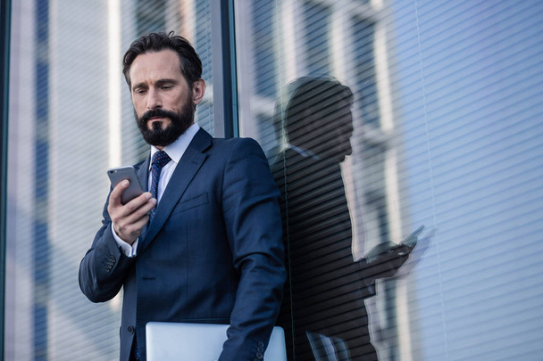 Constant communication. Concentrated bearded businessman using his smartphone while standing against glass wall - Φωτογραφία, εικόνα