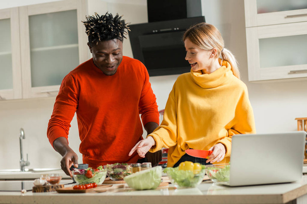 Hüftschwung eines glücklichen jungen internationalen Paares, das in der Küche steht und beim gemeinsamen Kochen auf Schüsseln mit Gemüse auf dem Tisch zeigt - Foto, Bild