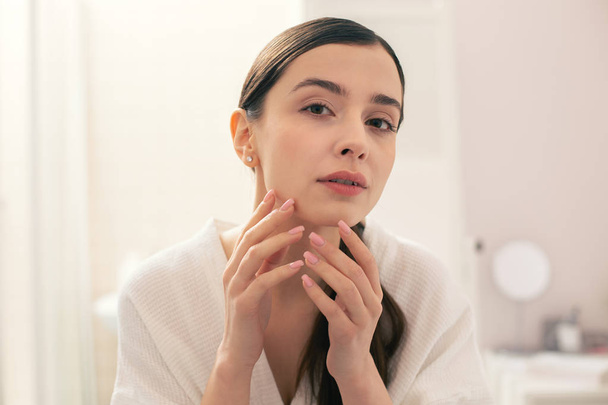 Calm young woman in a white towel looking concentrated while touching her chin and looking at the skin condition - Photo, Image