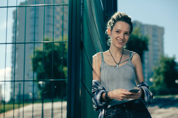 Positive young woman wearing comfortable casual clothes and smiling while standing with modern device next to the chain link fence - Photo, image