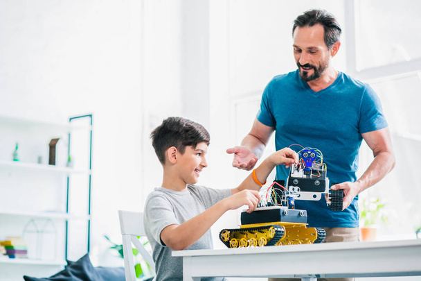 Hombre adulto alegre sosteniendo un dispositivo robótico mientras ayuda a su hijo con el proyecto de ingeniería
 - Foto, imagen