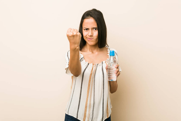 Jeune femme hispanique tenant une bouteille d'eau montrant poing à caméra, expression faciale agressive
. - Photo, image