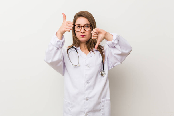 Νεαρή γιατρός γυναίκα σε ένα λευκό τοίχο δείχνοντας αντίχειρες πάνω και αντίχειρες κάτω, δύσκολη επιλογή έννοια - Φωτογραφία, εικόνα