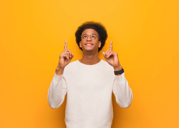 Jovem afro-americano sobre uma parede laranja surpreso apontando para cima para mostrar algo
 - Foto, Imagem