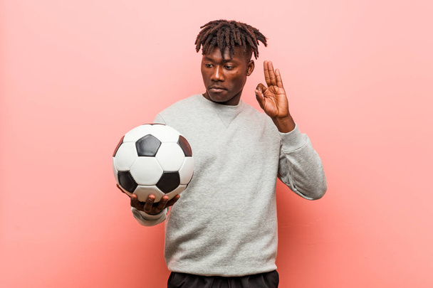 Fiatal fitness fekete ember, aki egy futball-labda vidám és magabiztos mutatja ok gesztus. - Fotó, kép