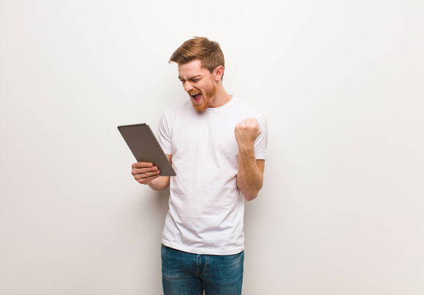 Young redhead man surprised and shocked. Holding a tablet. - Foto, afbeelding