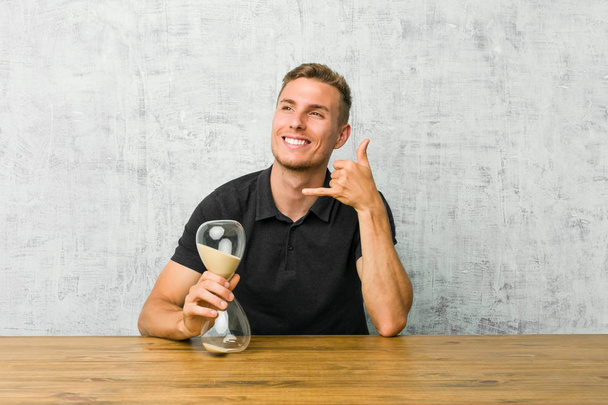 Young man holding a sand timer on a table showing a mobile phone call gesture with fingers. - Foto, Bild