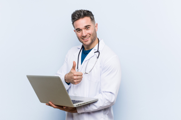 Kaukaski lekarz mężczyzna trzyma laptopa uśmiechnięty i podnoszenie kciuka w górę - Zdjęcie, obraz
