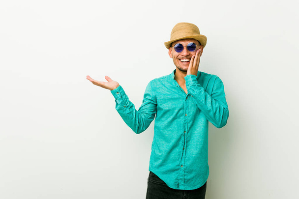 El joven hispano que usa ropa de verano tiene espacio para copiar en una palma, mantenga la mano sobre la mejilla. Sorprendido y encantado
. - Foto, Imagen