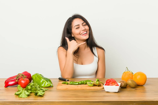 Young curvy woman preparing a healthy meal showing a mobile phone call gesture with fingers. - Φωτογραφία, εικόνα