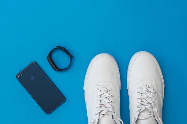 Blauwe Slimme armband, mobiele blauwe en witte sneakers met een blauwe achtergrond. Sport stijl. Platte lay. - Foto, afbeelding