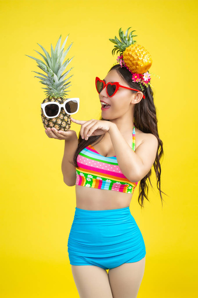 Beautiful woman in a swimsuit holding a pineapple poses on a yellow background. - Foto, Imagen