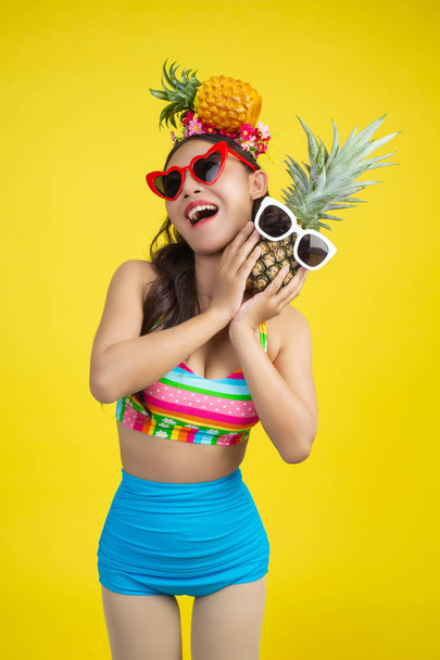 Beautiful woman in a swimsuit holding a pineapple poses on a yellow background. - Фото, изображение