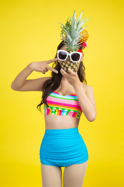 Beautiful woman in a swimsuit holding a pineapple poses on a yellow background. - Valokuva, kuva