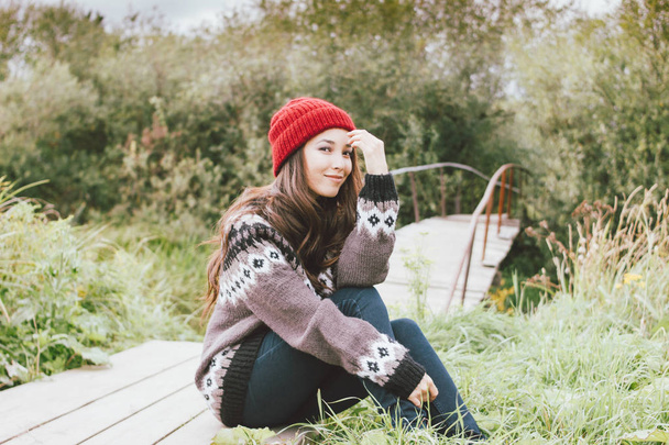 Beautiful carefree long hair asian girl in the red hat and knitted nordic sweater sitting in autumn nature park, travel adventure lifestyle - Photo, Image
