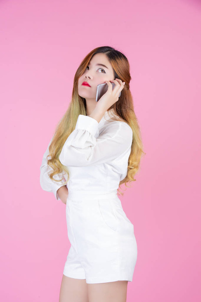 A beautiful woman dressed with a white dress, showing the phone and facial emotions on a pink background. - Valokuva, kuva