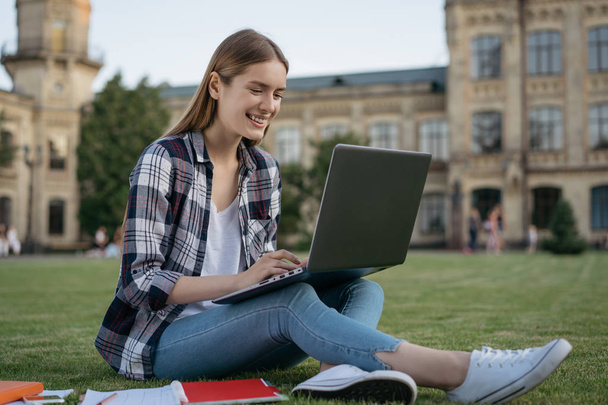 Attractive smiling woman using laptop computer, searching information, typing on keyboard, working project online. Successful university student studying, learning language sitting on grass in park. Online education concept - Foto, imagen
